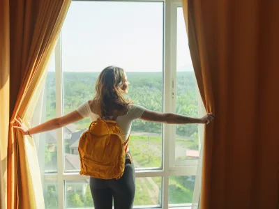 Eine Frau, die einen Rucksack trägt, steht vor einem Fenster und öffnet den Vorhang mit beiden Händen.