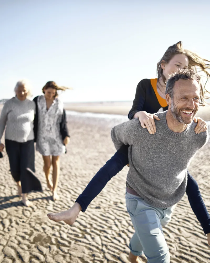 Eine Familie läuft gemeinsam am Strand entlang. Der Fokus liegt auf einem Mann, der ein Mädchen auf dem Rücken trägt.